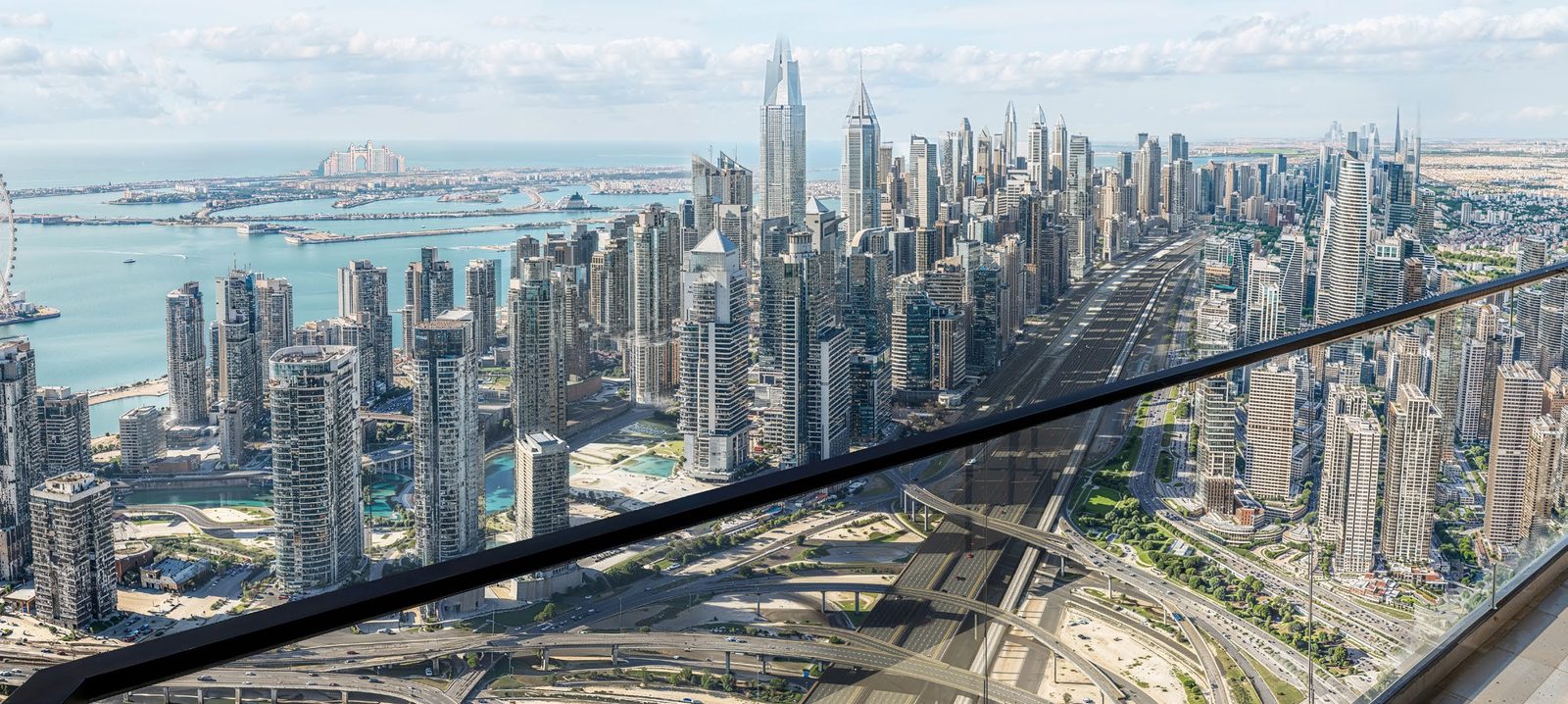 Dubai Marina and the sea seen from high above Sheikh Zayed Road, behind a glass balustrade.